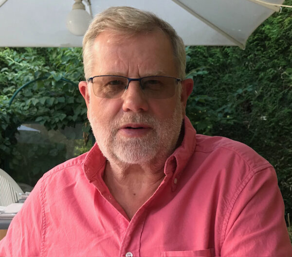 A head and shoulders shot of a older bearded white man wearing a pink shirt and glasses.