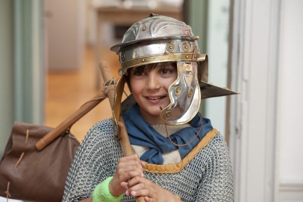A boy dressed in a Legionary Soliders uniform with a helmet and chainmail and holding his Sacrina (marching pack)