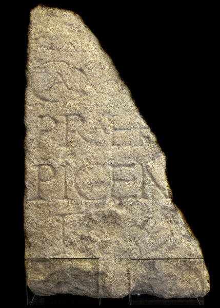 An irregular half of a carved stone slab with incomplete text carved in large letters covering most of it's face