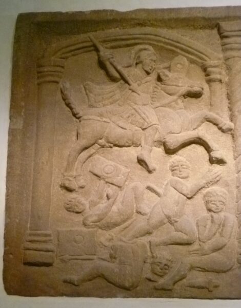 A Roman carved stone showing a horse and rider jumping over a variety of people who appear to have been in a battle, as well as various shields and weapons