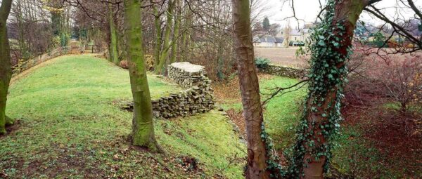 A photo taken from the top of a ramparts, with some immature trees scattered upon it, and looking towards a defensive ditch