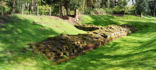 A photo showing an 'L' shaped base of a sustantial stone wall, with grass banks on either side, and tree cover to the left hand side