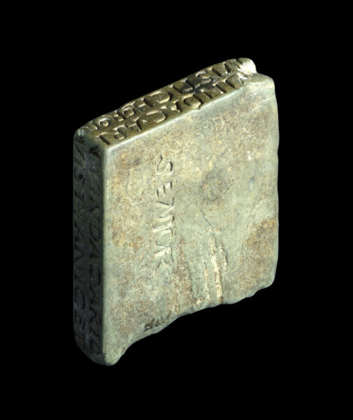 An irregular small stone square collyrium stamp (looks like a thick tile) - this stone is shown at an oblique angle to show carved text along two edges - there is also text carved onto the face of this square