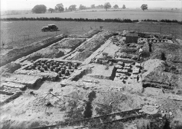 A black and white photo showing a complex of archaeological features - there is also a car parked nearby which gives the photo a reference to its 1930's origin - also in the foreground there is also the end of a narrow gauge railway track