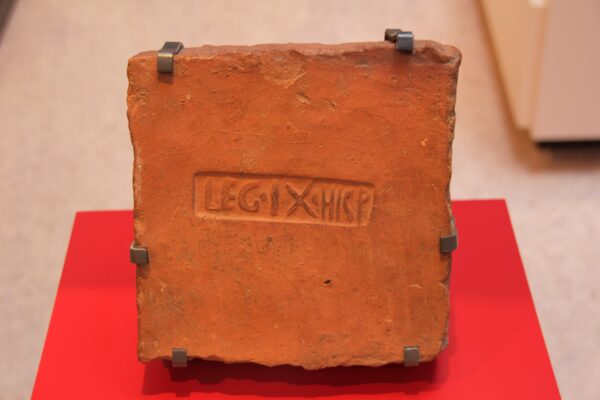 A colour photo of a terracotta roof tile presented on a metal display stand which is clasped in several places along the tiles edge