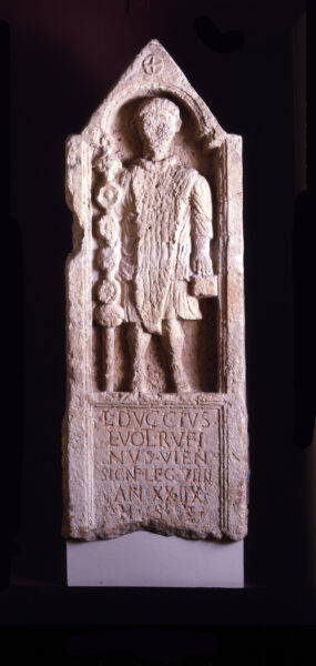 A colour image showing an almost complete carved tombstone of Lucius Duccius Rufinus wearing robes - he is holding his standard in his right hand, and a square object in his left hand - he is standing within an arched alcove with a double stepped impost, and a triangular apex that is decorated with a cross within a circle - below this is a carved inscription - both these have a double inset border