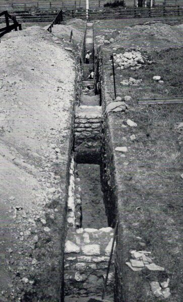 A black and white photo showing a neatly dug, deep trench containing substantial stone walls that intersect this trench - there is an extensive spoil heap on the left hand side, and much small collections of stone of the right hand side - there is also a measuring sticks in the foreground, and another about halfway along the trench - three men can be seen within the trenches, and in the background there is a fence-line along either side of the railway line