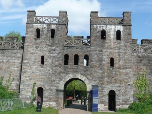 A colour photo showing a large stone built gatehouse with a main entrance gate, plus two much smaller side gates - above this there is a level with three central windows, and two slightly smaller window that mirror the positions as described previously - above this is another level that links to a palisade walkway - and finally above this are two towers on either side of the main gate with another platform placed at the top of each tower - there is also a variety of street furniture such as litter bins, fences and signage