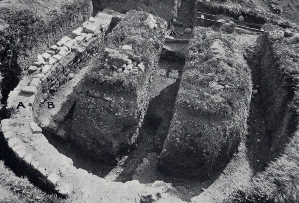 A black and white photo showing the horseshoe shaped wall of an apsidal building and within this is a rectangular shaped earlier building - within this are two mounds of earth with a neatly cut trench running through it - there are a couple of pieces of archaeological equipment on the top left hand side that look like a folded up canvas and a wooden framework