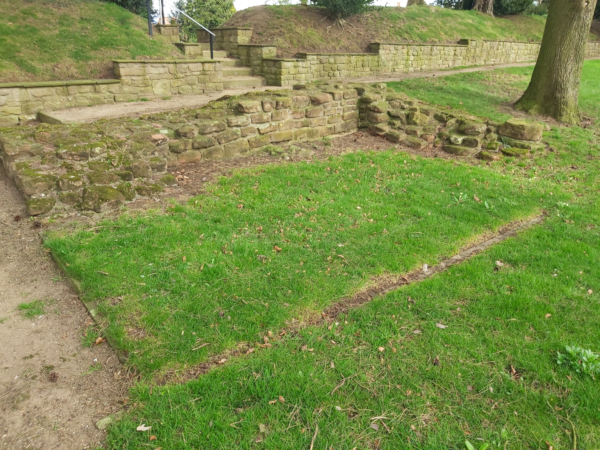 A colour photo showing two interescting walls about five courses high, with an outline on the other two sides of a rectangular area of grass - There is a pathway on the left hand side that turns right along the longest section of wall, and then follows a modern wall that skirts the edge of a grass embankment - there are also steps leading throught this embankment - mature trees are visable in the background