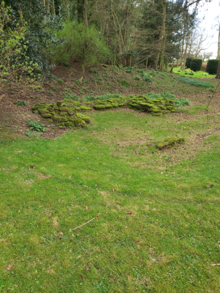 A colour photo showing parts of the outline of the corner tower, which are bright green with moss cover - the area is undulating, with a graas bank in the foreground, and rougher ground leading to mature trees in the background - in the far top right, well maintained yew hedging can be seen