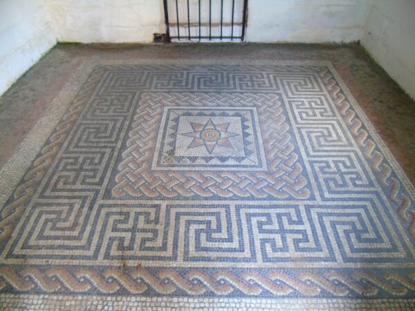 A colour photo showing a Roman mosaic floor - this mosaic has a central star or sun shape within a decorated square - surrounding this are three wide borders of tesserae - this room has white washed walls, and the base of an iron grating is visable against the back wall of this room