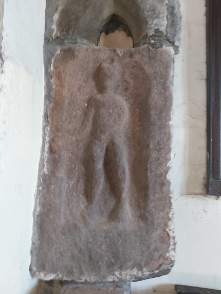 A colour photo showing a well worn stone carving of the Roman god Mercury - this stone has a red hue to it, and above this is a gothis shaped arch which doesn't seem to match up with this carving