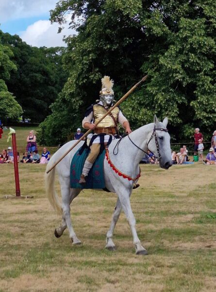 A colour photo of a Roman solider riding a white horse across orchard fields in Malton, and holding a spear (pilum)