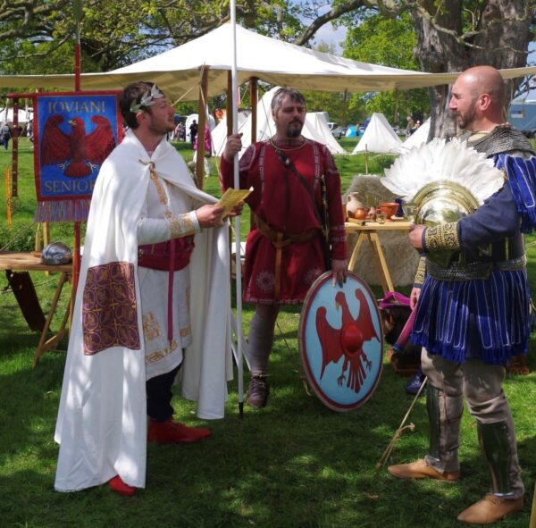 A colour photo showing a declaration being read to a soldier holding a helmet, by a white robed Roman - another Roman soldier stands guard in the background