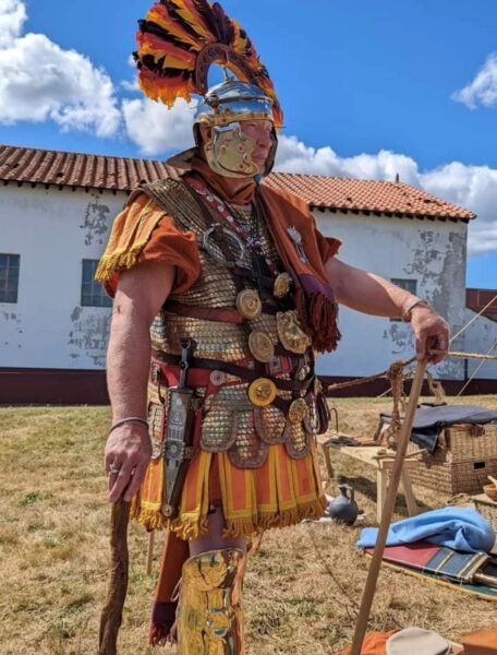 A colour photo of a Roman Soldier standing and ready for battle - there are also various pieces of equipment in the background