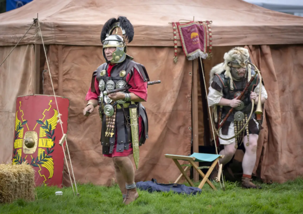 A colour photo showing one member of Legio VI stepping forward well armed, and holding his pugio (dagger) whilst another is leaving their tent holding a horn