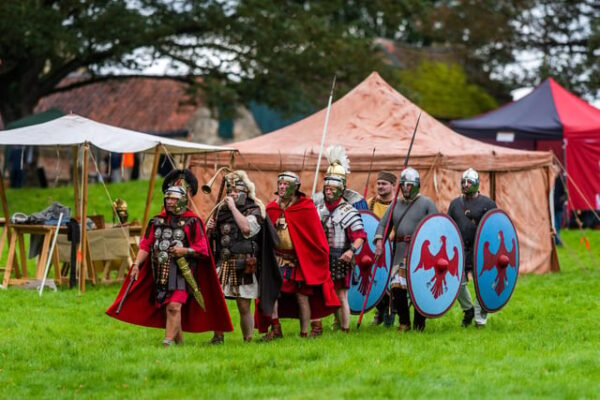 A colour photo showing eight Roman soldiers marching from right to left alongside their camp - the second soldier is blowing into a large horn