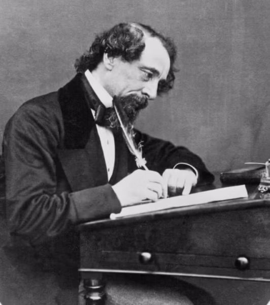 A black and white photograph of the author Charles Dickens writing at a desk using a feather quill.