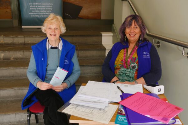 Two female volunteers seated at the entrance to a museum, both smiling.