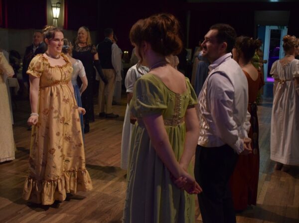 Men and women in Georgian period costume stand facing each other, about to begin a formal dance.