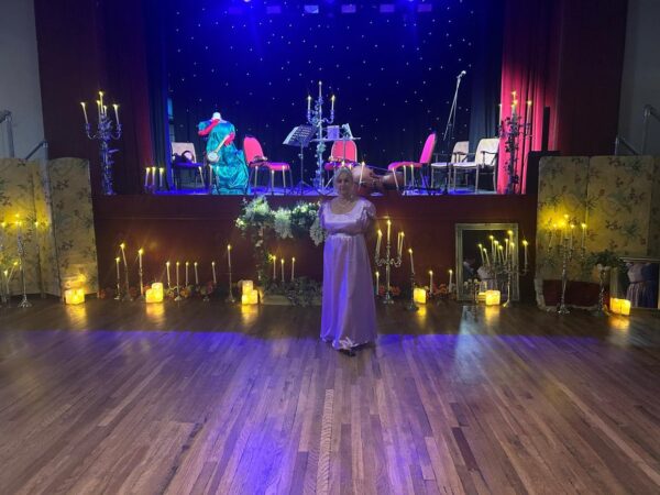 A woman in a lilac Georgian dress stands in front of a stage decorated with candles.