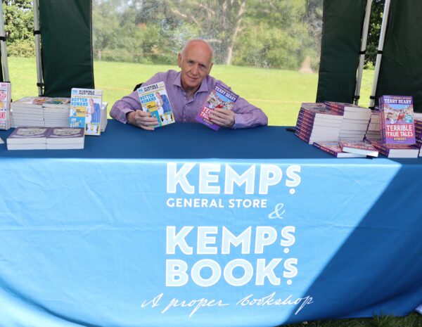 Author Terry Deary holds up copies of his book. He sits behind a blue table in a tent in a field in Malton, North Yorkshire.