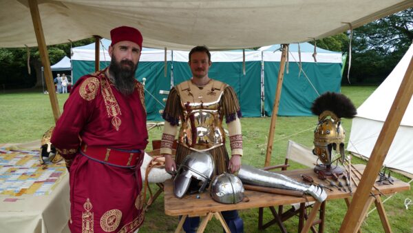 Two men stand, one dressed in gold Roman armour, the other in red civilian Roman attire