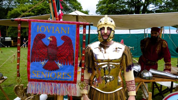 Man dressed as Roman soldier in gold armour stands next to banner with red bird